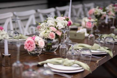 A large, long, decorated, wooden table and chairs, covered with a white tablecloth with dishes, flowers, candles.