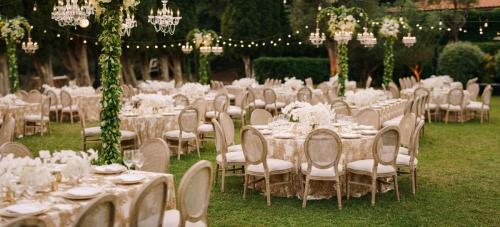 Wedding dinner table reception. Elegant tables for guests with cream tablecloths with patterns, on green lawn, with garlands and chandeliers hanging over them. Chairs with round back